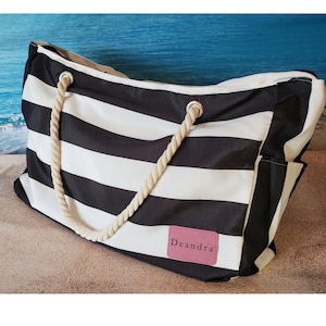 May include: A large black and white striped tote bag with rope handles. The bag has a pink rectangular patch with the name "Deandra" on it. The bag is sitting on a sandy surface with a blue ocean background.