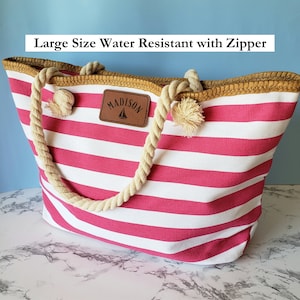 May include: A large pink and white striped tote bag with a brown leather patch that says "MADISON". The bag has rope handles and a zipper closure. It is water resistant.