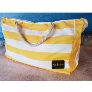 May include: Yellow and white striped beach tote bag with rope handles. The bag has a rectangular shape and a brown leather patch with the name "Kristy" on it. The background shows a beach scene.