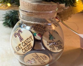 Op de afbeelding: Een heldere glazen pot met een kurken deksel, gevuld met houten schijfjes. De schijfjes hebben feestelijke tekst en illustraties, waaronder "EAT CANDY CANES" en een zuurstok, en "DECORATE THE TREE" met een kerstboom. De pot is vastgebonden met touw.