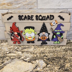 May include: Wooden Halloween sign with the words "SCARE SQUAD" and four cartoon characters: a devil, Frankenstein's monster, a gnome with a pumpkin, and a witch. The sign includes bats and a candy corn.