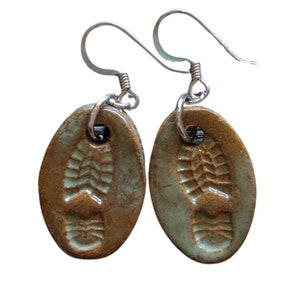 May include: A pair of ceramic earrings with a green and brown glaze. The earrings are shaped like footprints and have silver hooks.