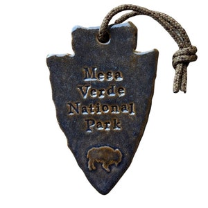 May include: A brown ceramic arrowhead ornament with a brown leather cord. The arrowhead has the text "Mesa Verde National Park" and a silhouette of a bison.