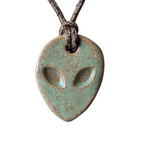 May include: A teal ceramic alien pendant with a brown cord. The pendant features a textured surface and two oval eye shapes. The cord is a twisted brown color, knotted at the top.