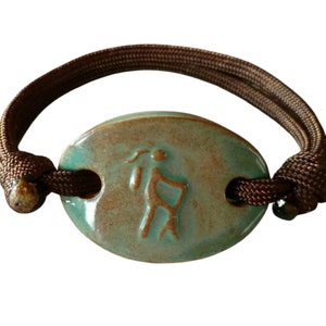 May include: A green ceramic bracelet with a brown cord. The ceramic charm features a stylized figure of a person.