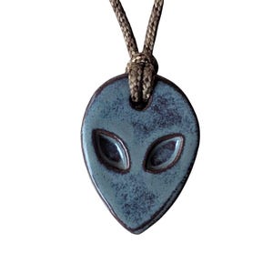 May include: A blue-green ceramic alien pendant with a brown cord. The pendant features a stylized alien head with large, oval eyes. The cord is knotted at the top, and the pendant has a glossy finish.