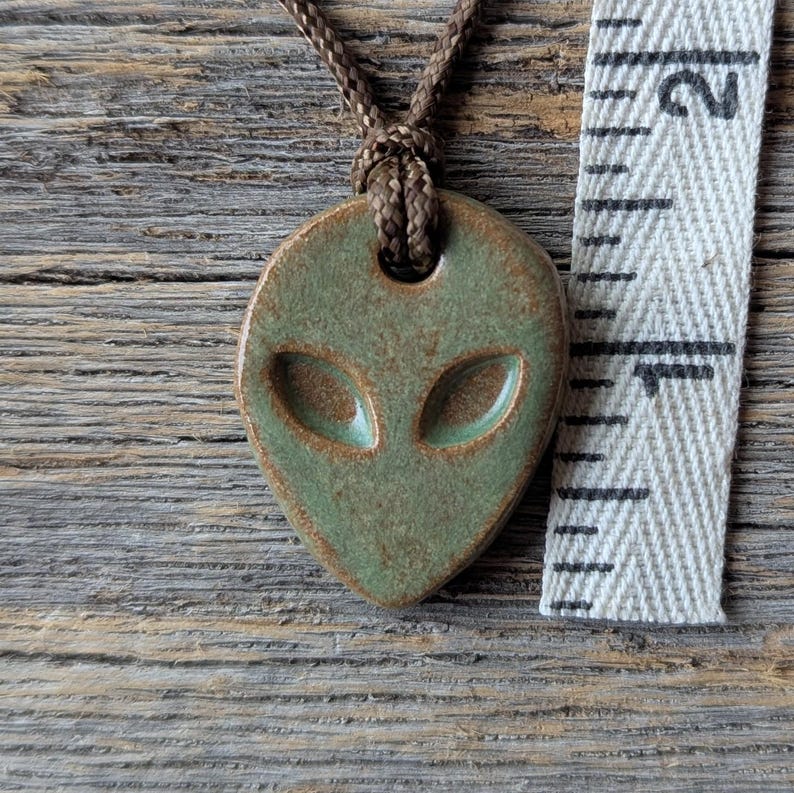 以下が含まれることがあります： 緑と茶色のセラミックエイリアンペンダント。茶色のコードで吊り下げられています。ペンダントは、大きな楕円形の目を持つ様式化されたエイリアンの顔が特徴です。メジャーテープはペンダントのサイズを示しています。