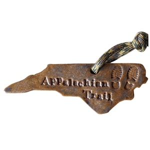 May include: A brown metal keychain shaped like the state of North Carolina with the words "Appalachian Trail" and two footprints embossed on it. The keychain is attached to a green and yellow paracord.
