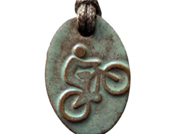 May include: Oval pendant with a cyclist design. The pendant has a distressed, turquoise-green finish with a brown bicycle and rider silhouette. It hangs from a brown cord, creating a rustic, outdoorsy aesthetic. The pendant is suitable for cyclists.