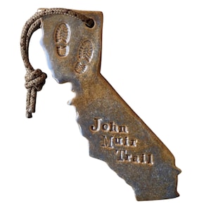May include: A brown, ceramic ornament shaped like the state of California with the words "John Muir Trail" inscribed on it. The ornament has a brown rope attached to the top for hanging.