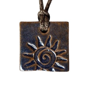 May include: Square pendant with a sun design. The pendant is a dark blue-brown color with a textured surface. The sun design is carved into the pendant and is a lighter brown color. The pendant is attached to a brown cord.