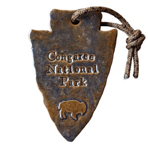 May include: A brown ceramic arrowhead ornament with a tan rope attached. The arrowhead has the text "Congaree National Park" and a silhouette of a buffalo.