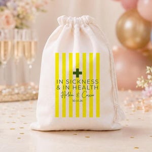 May include: A cream-colored drawstring pouch featuring a vertical yellow striped pattern. The center displays a dark green cross icon above the text IN SICKNESS & IN HEALTH, followed by the names Holden & Cassie and the date 10.10.26.