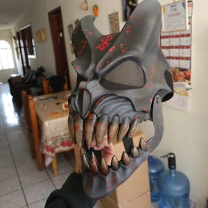 Könnte beinhalten: Eine schwarze Totenkopfmaske mit gezackten Zähnen und roten Spritzern. Die Maske hat ein bedrohliches Design mit großen, spitzen Zähnen und einem Symbol auf der Stirn. Die Maske wird von einer Person gehalten.