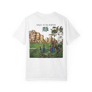 以下が含まれることがあります： 緑豊かな風景の前で立っている人々のグループが描かれたレゲエアルバムのカバーのグラフィックプリントが施された白いTシャツ。アルバムのタイトルは「Tribute to the Martyrs」、バンド名は「Steel Pulse」。