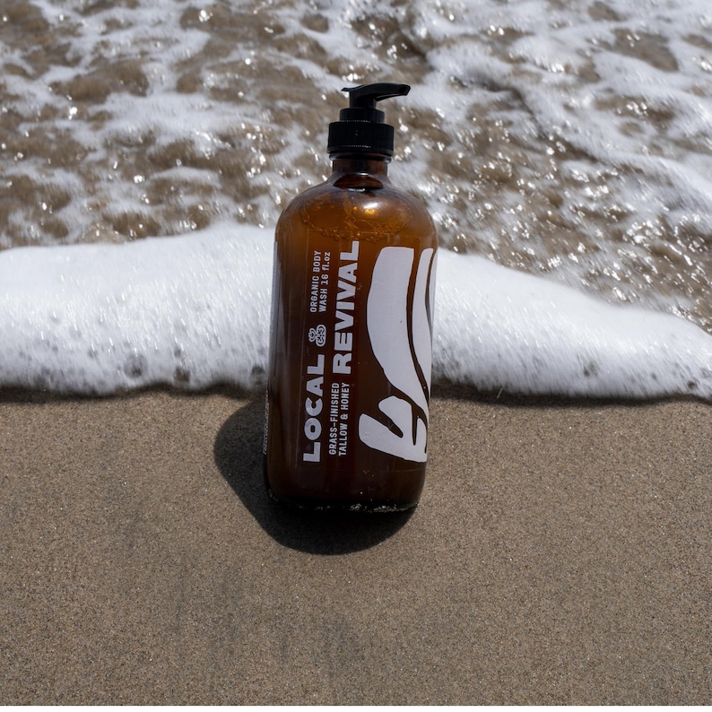 May include: A brown glass bottle of "Local Revival" organic body wash with a black pump dispenser. The label features white text and a leaf design. The bottle is on a sandy beach with ocean waves in the background.