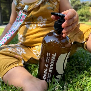 May include: A brown bottle of organic body wash with a black pump, held by a child. The bottle has white text that reads "LOCAL & REVIVAL" and "grass-finished tallow & honey". The child is wearing a mustard yellow romper and sitting on grass.