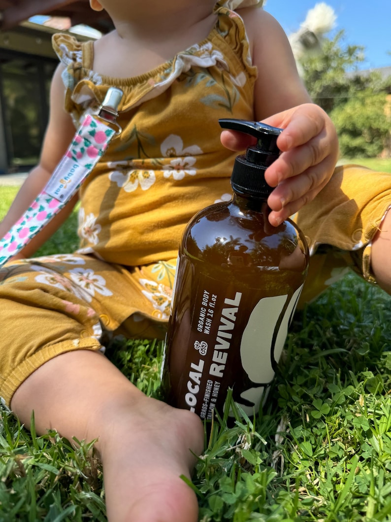 May include: A brown bottle of organic body wash with a black pump, held by a child. The bottle has white text that reads "LOCAL & REVIVAL" and "grass-finished tallow & honey". The child is wearing a mustard yellow romper and sitting on grass.