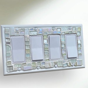 May include: White light switch plate with four toggle switches, decorated with a mosaic of iridescent white and light green glass tiles.