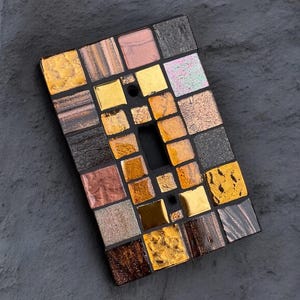 May include: Rectangular mosaic tile with a central rectangular opening. The mosaic is composed of small, square tiles in shades of gold, brown, black, and iridescent colors. The tile is set against a textured, gray background.