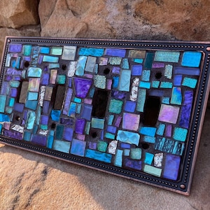 May include: A decorative light switch cover with a mosaic design featuring blue, purple, and white glass tiles. The cover has a copper-colored frame with a beaded edge.