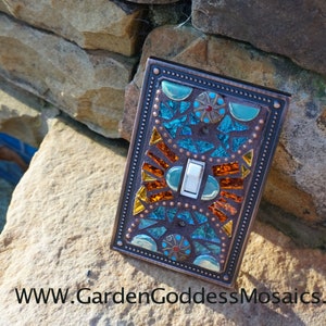 Pu&ograve; includere: Una placca per interruttore con un design a mosaico in tonalit&agrave; turchese, arancio e rame. Il mosaico &egrave; circondato da una cornice color rame con un bordo decorativo.