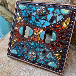 May include: A double light switch cover with a mosaic design featuring turquoise, orange, and gold glass tiles. The cover has a copper-colored frame with a beaded border.