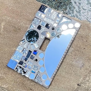 May include: A rectangular light switch cover plate decorated with a mosaic of various glass pieces, including small blue, black, and clear tiles, a circular mirror, and a large crescent-shaped mirror piece, all set into a light gray grout base.
