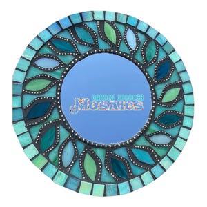 May include: A round mosaic mirror featuring a central reflective surface with the text Garden Goddess Mosaics. The frame is composed of teal, light blue, and green glass pieces arranged in a leaf-like pattern with dark grout lines throughout.