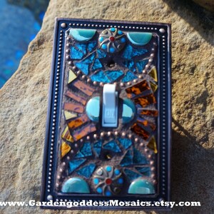 Pu&ograve; includere: Una placca per interruttore della luce in mosaico con uno schema di colori turchese, arancione e marrone. La placca dell'interruttore ha una cornice metallica decorativa con un bordo perlato. L'interruttore &egrave; bianco e dice "ON" in nero.