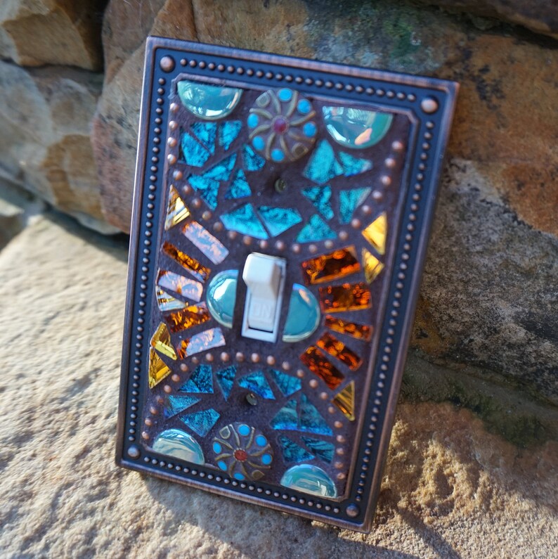 Pu&ograve; includere: Una placca per interruttore della luce in rame con un design a mosaico con piastrelle di vetro blu, verde, arancio e marrone. L'interruttore &egrave; bianco.