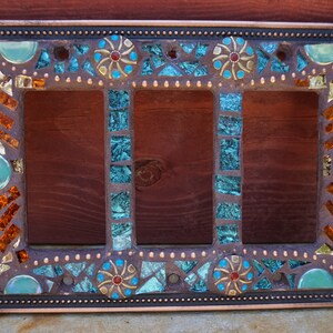 Pu&ograve; includere: Una placca di copertura per interruttore triplo con un design a mosaico. Il mosaico presenta piastrelle di vetro turchese, blu, oro e arancio. La placca &egrave; in metallo e ha una finitura in rame.