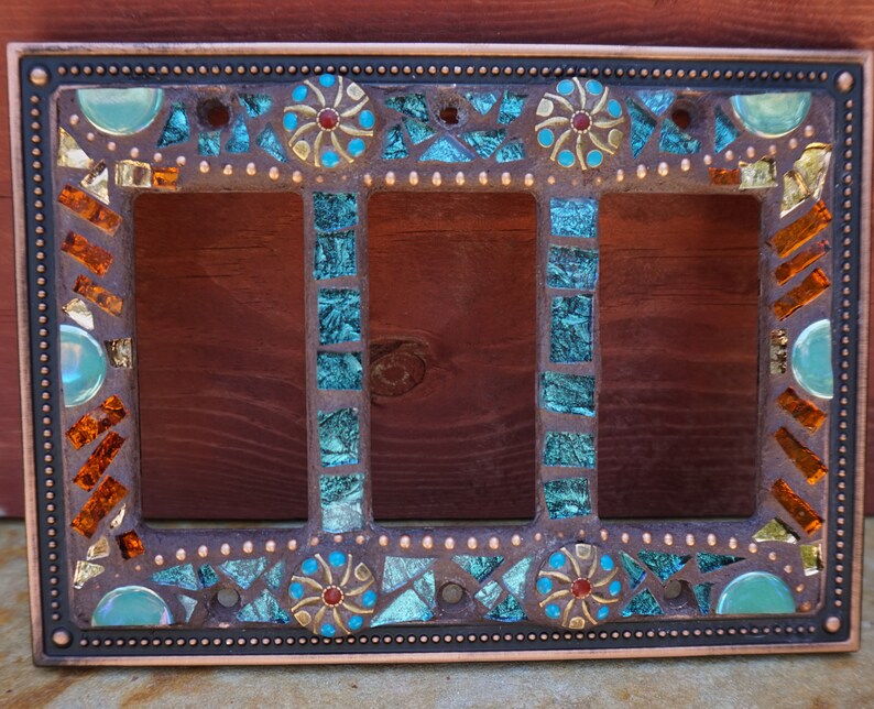 Pu&ograve; includere: Una placca di copertura per interruttore triplo con un design a mosaico. Il mosaico presenta piastrelle di vetro turchese, blu, oro e arancio. La placca &egrave; in metallo e ha una finitura in rame.