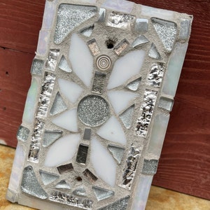 Puede incluir: Un azulejo de mosaico blanco y plateado con un dise&ntilde;o floral. La flor tiene cinco p&eacute;talos y un c&iacute;rculo central. El azulejo est&aacute; hecho de peque&ntilde;os trozos de vidrio y est&aacute; colocado en un mortero gris.