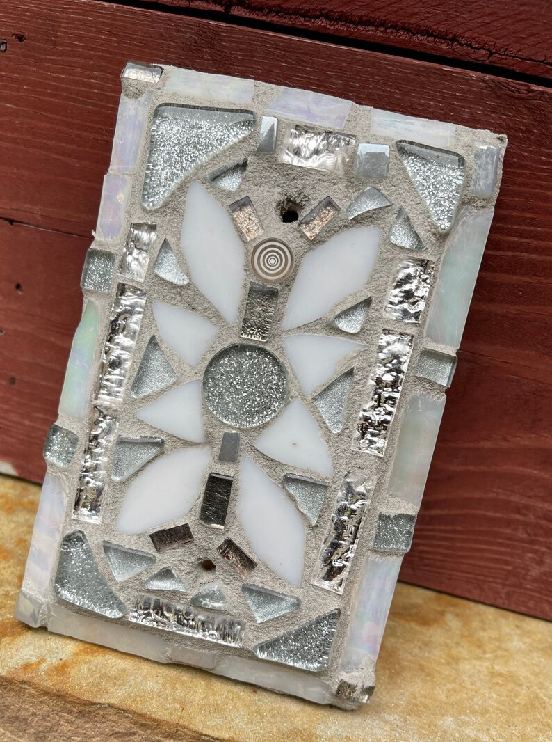 Puede incluir: Un azulejo de mosaico blanco y plateado con un dise&ntilde;o floral. La flor tiene cinco p&eacute;talos y un c&iacute;rculo central. El azulejo est&aacute; hecho de peque&ntilde;os trozos de vidrio y est&aacute; colocado en un mortero gris.