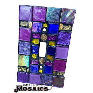 May include: A rectangular light switch cover plate decorated with a mosaic of square glass tiles in shades of purple, blue, and yellow, featuring iridescent finishes and small gold-toned circular accents with spiral patterns embedded throughout.