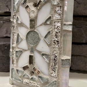 Puede incluir: Un azulejo de mosaico blanco y plateado con un dise&ntilde;o floral. El azulejo est&aacute; hecho de peque&ntilde;os trozos de vidrio y est&aacute; colocado en un mortero gris.