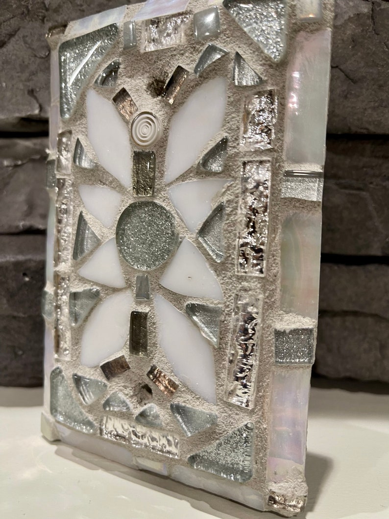 Puede incluir: Un azulejo de mosaico blanco y plateado con un dise&ntilde;o floral. El azulejo est&aacute; hecho de peque&ntilde;os trozos de vidrio y est&aacute; colocado en un mortero gris.