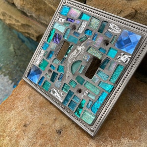 May include: A light switch cover with a mosaic design featuring blue, green, and silver glass tiles. The cover has a silver metal frame with a beaded edge.
