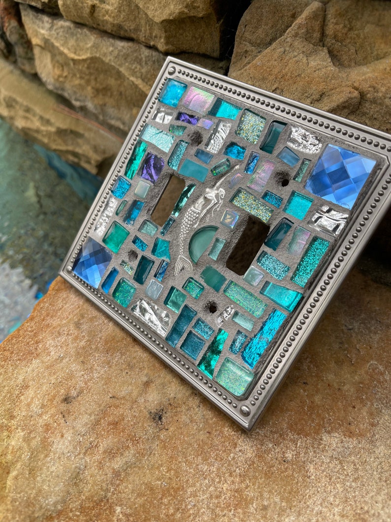 May include: A light switch cover with a mosaic design featuring blue, green, and silver glass tiles. The cover has a silver metal frame with a beaded edge.