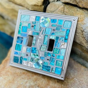 May include: A light switch cover with a mosaic design featuring blue and green glass tiles, a mermaid, and a crystal. The cover has a silver metal frame with a beaded border.