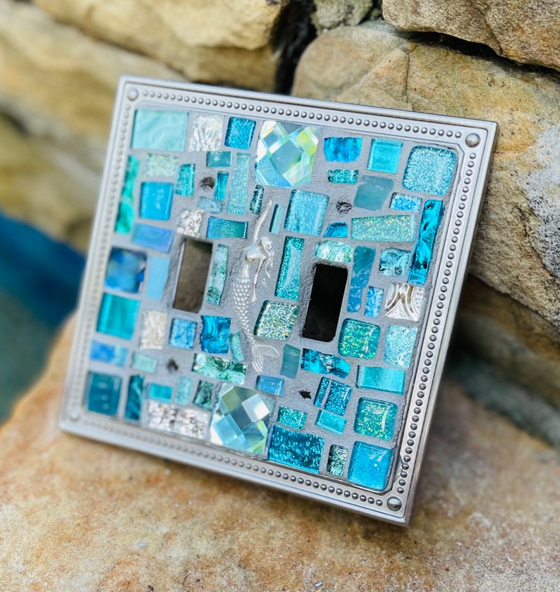 May include: A light switch cover with a mosaic design featuring blue and green glass tiles, a mermaid, and a crystal. The cover has a silver metal frame with a beaded border.