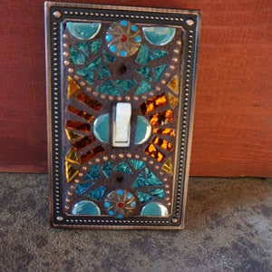 Pu&ograve; includere: Una placca per interruttore della luce in rame con un design a mosaico con piastrelle di vetro turchese, arancio e oro. Il design a mosaico include un motivo circolare con un centro rosso e dorato.