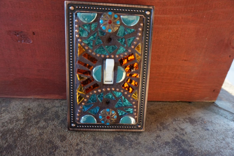 Pu&ograve; includere: Una placca per interruttore della luce in rame con un design a mosaico con piastrelle di vetro turchese, arancio e oro. Il design a mosaico include un motivo circolare con un centro rosso e dorato.
