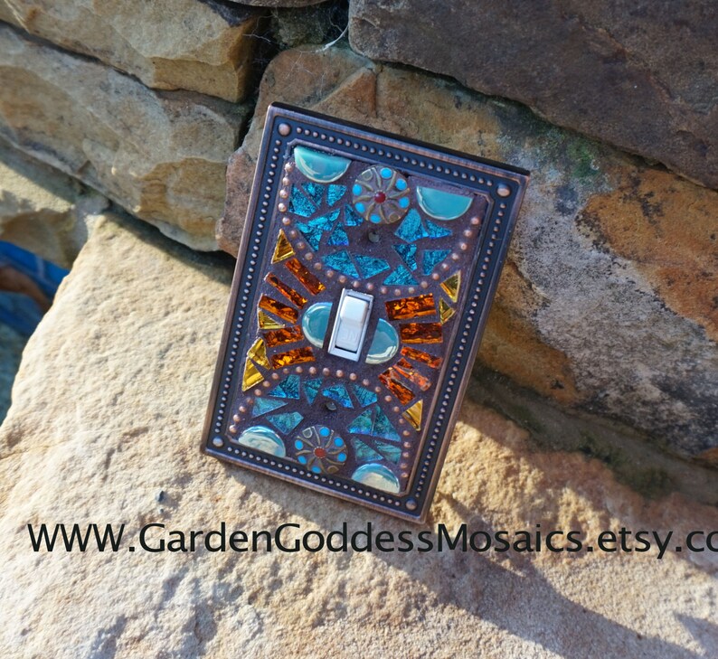 Pu&ograve; includere: Una placca per interruttore con un design a mosaico in tonalit&agrave; turchese, arancio e rame. Il mosaico &egrave; circondato da una cornice color rame con un bordo decorativo.