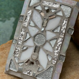 Puede incluir: Un azulejo de mosaico blanco y plateado con un dise&ntilde;o geom&eacute;trico. El azulejo est&aacute; hecho de peque&ntilde;os trozos de vidrio y est&aacute; colocado en un mortero gris.