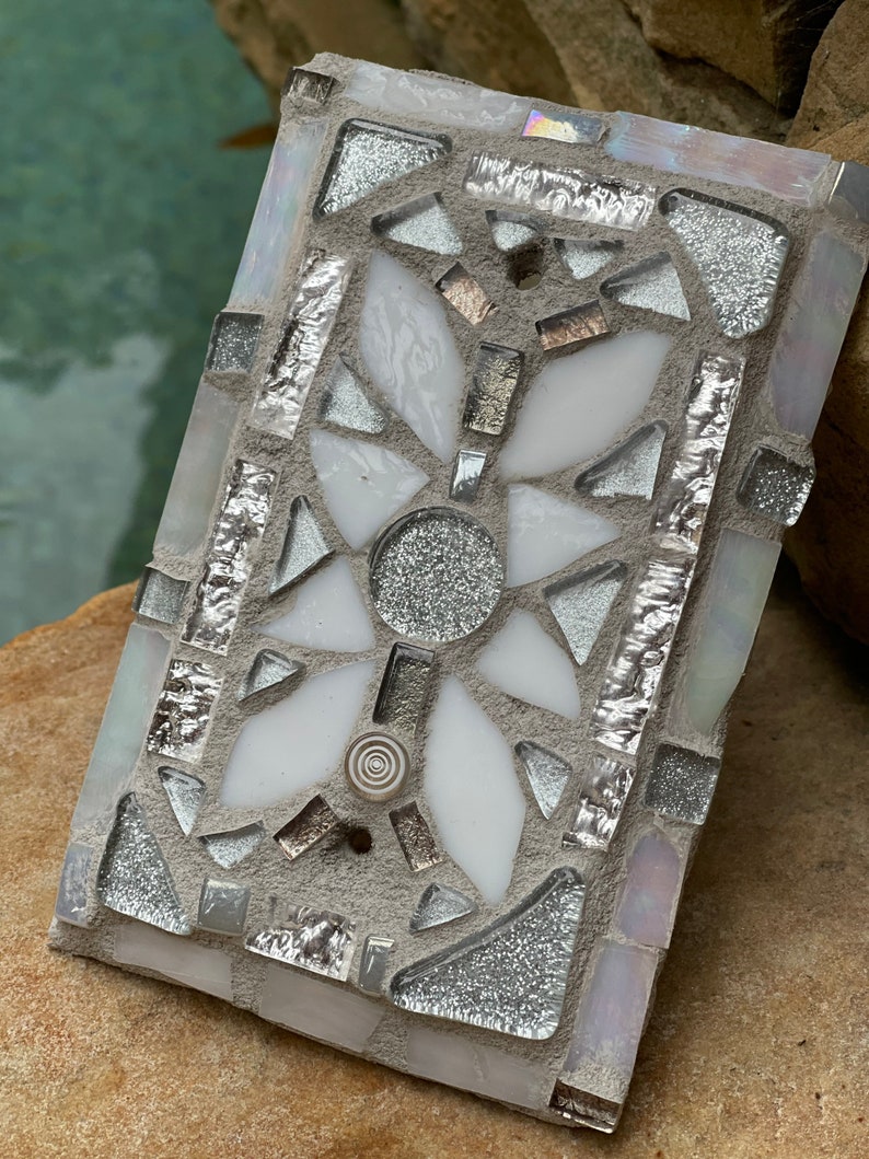 Puede incluir: Un azulejo de mosaico blanco y plateado con un dise&ntilde;o geom&eacute;trico. El azulejo est&aacute; hecho de peque&ntilde;os trozos de vidrio y est&aacute; colocado en un mortero gris.