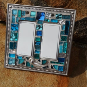 May include: A double light switch cover with a mosaic design featuring blue and green glass tiles. A mermaid design is incorporated into the mosaic. The cover has a silver metal frame.