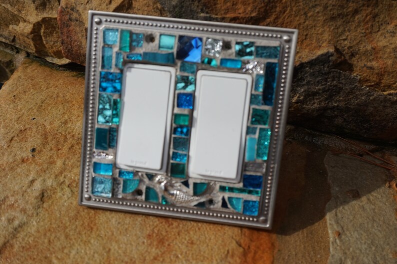 May include: A double light switch cover with a mosaic design featuring blue and green glass tiles. A mermaid design is incorporated into the mosaic. The cover has a silver metal frame.