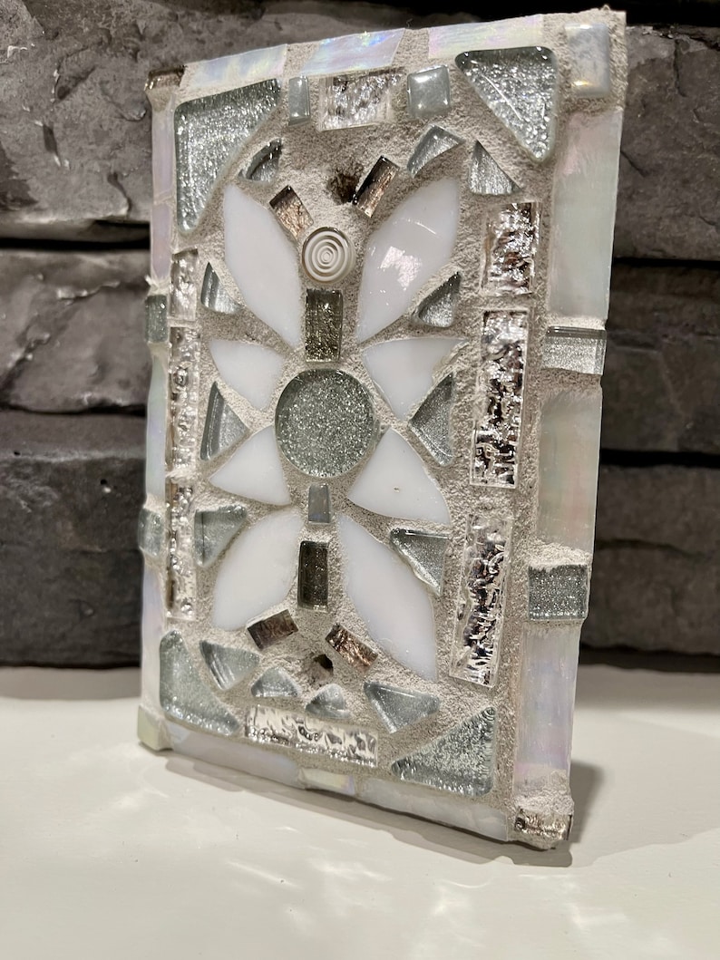 Puede incluir: Un azulejo de mosaico blanco y plateado con un dise&ntilde;o geom&eacute;trico. El azulejo est&aacute; hecho de peque&ntilde;os trozos de vidrio y est&aacute; colocado en un mortero blanco.